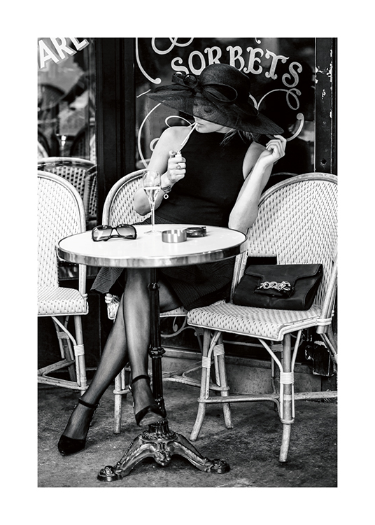  – Black and white photograph of a woman sitting outside of a café, wearing a hat and lighting a cigarette